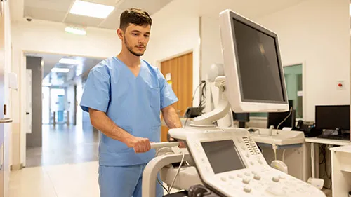 A sonographer wheels a sonography cart through a hospital hallway.