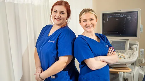 Two female graduates from Concorde's Diagnostic Medical Sonography Program in blue scrubs smiling.