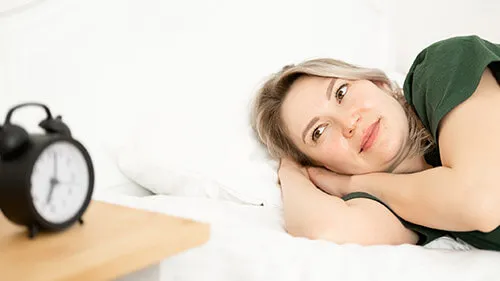A woman in bed smiles slightly as she looks at the alarm clock on her side table.
