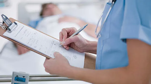 A health care professional takes notes on a clipboard while a patient lies on a hospital bed in the background.