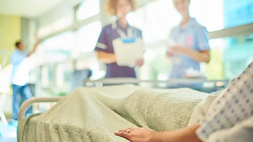 Close-up of a patient's hand as they rest in a hospital bed. Two health care professionals look on in the background.