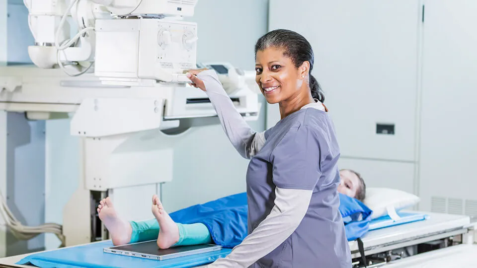A smiling woman wearing scrubs positions X-ray equipment above a young girl's foot.