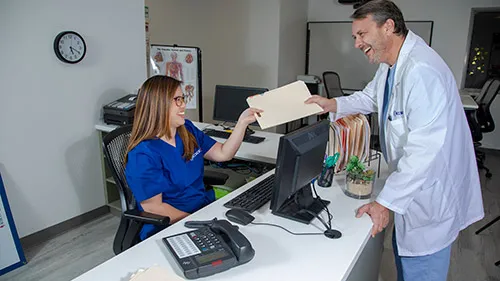 Medical office professional behind desk hands chart to doctor.