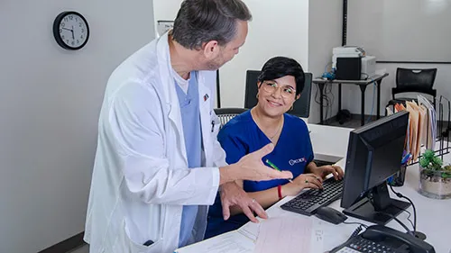 A smiling medical office professional listens to doctor while at her desk.